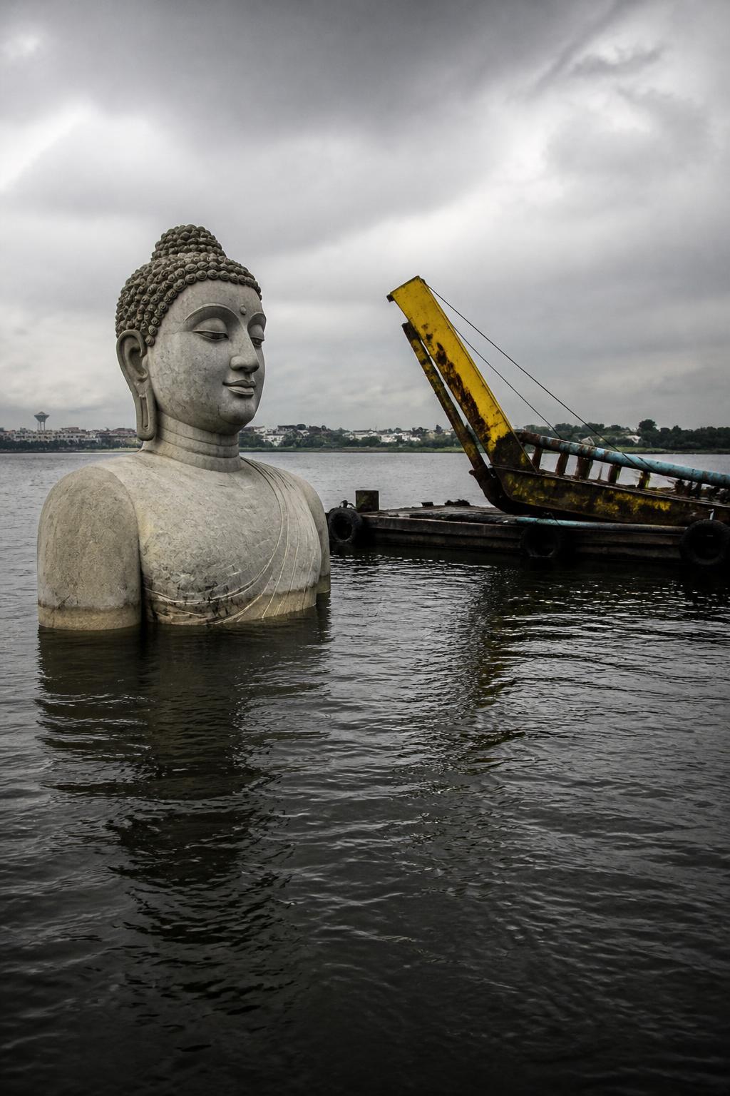 A massive Buddha statue partially submerged beside a barge on a lake.